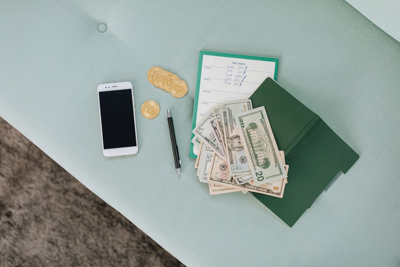 Flat lay of a smartphone, US dollar bills, coins, and a planner on a green surface.