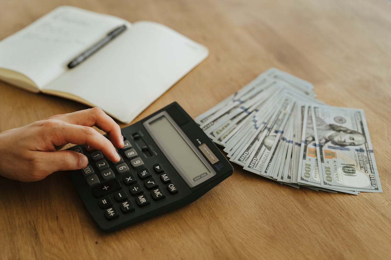 Close-up of a hand using a calculator with cash and a notebook on a wooden table.